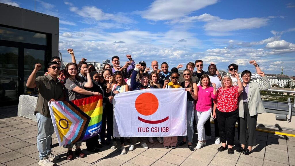 The same group of international unionists standing on the roof deck of a building. Clouds are in the background and everyone has their fists raised in a symbol of solidarity.