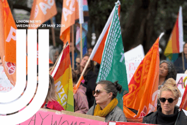 A crowd at an outdoor rally holding orange union flags, rainbow Pride flags, PSA flags, and a green and white koru-design flag. In the foreground, two women are seated — one in sunglasses and a mustard-yellow scarf, the other in dark clothing and round sunglasses. A pink placard is held up at the front. The "Union News, Wednesday 27 May 2026" masthead and CTU logo appear in the top-left corner.