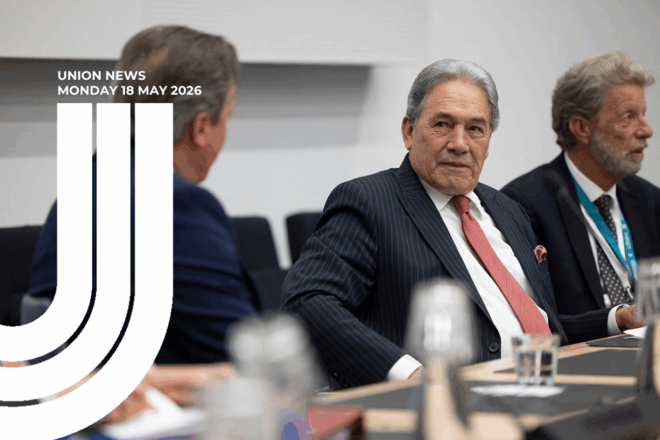 A photograph of NZ First leader Rt Hon Winston Peters seated at a boardroom-style meeting table. He is wearing a dark pinstriped suit, white shirt and red tie with a red pocket square, and is looking towards the camera with a slight smile. To his left (viewer's right) is an older man in a dark suit and patterned tie wearing a lanyard, looking off to the side. In the foreground on the left, slightly out of focus, is the back of another man's head and shoulders in a navy jacket. Water glasses, a bottle and papers sit on the wooden table in front of them. The setting is a modern meeting room with pale grey walls. Overlaid on the left side of the image is a white "Union News — Monday 18 May 2026" masthead with a stylised half of a U logo beneath it