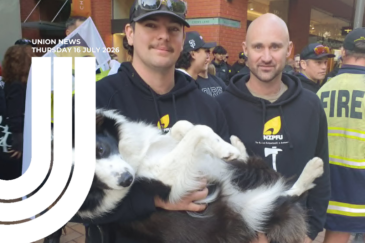 Two firefighters in black NZPFU hoodies smile at the camera at a rally, one is holding a black and white border collie on its back. Behind them, other firefighters gather in a city laneway, including one in high-vis fire kit. Text overlay reads "Union News, Thursday 16 July 2026".