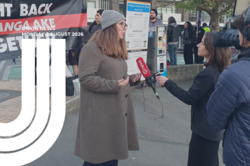 Melissa Ansell-Bridges wearing a grey beanie and brown coat speaks to a 1News reporter, with picketing workers and a union banner behind her.