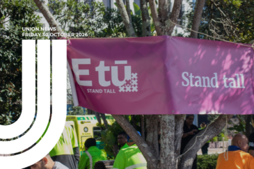 Graphic for Union News: Friday 30 October 2026. Colour photo of a pink E tū banner reading “Stand tall” strung between trees at an outdoor rally. Below it, people in high-visibility workwear stand around with an ambulance parked behind them. White curved lines overlay the left side of the image.